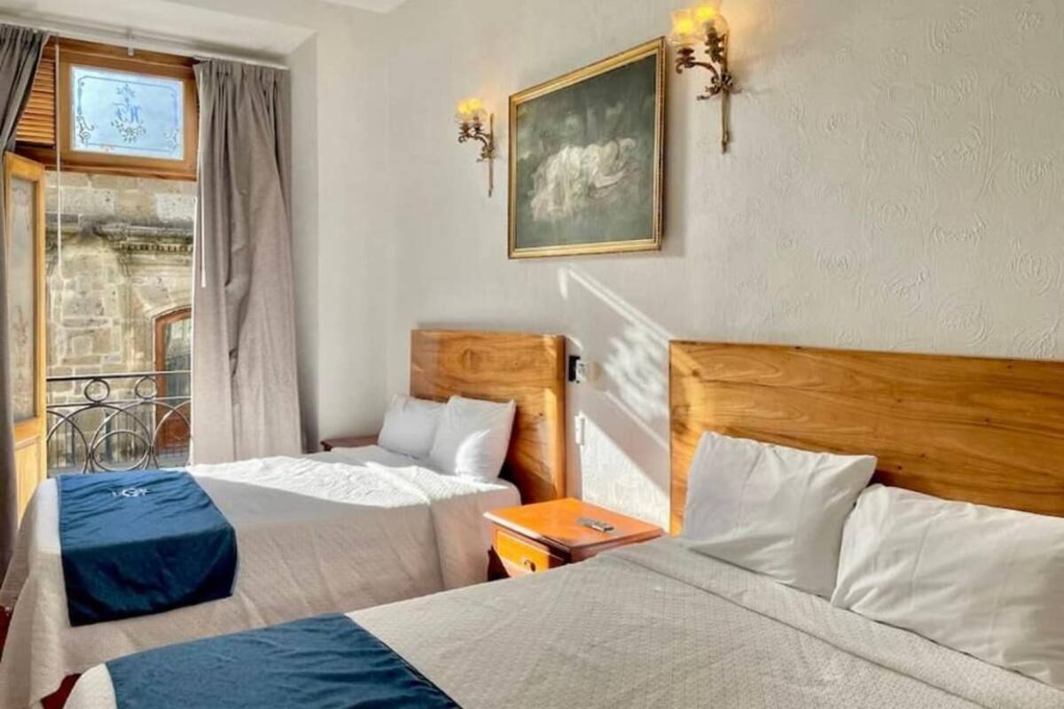 A hotel room with two beds, a wooden headboard, a painting on the wall, and a window with curtains.