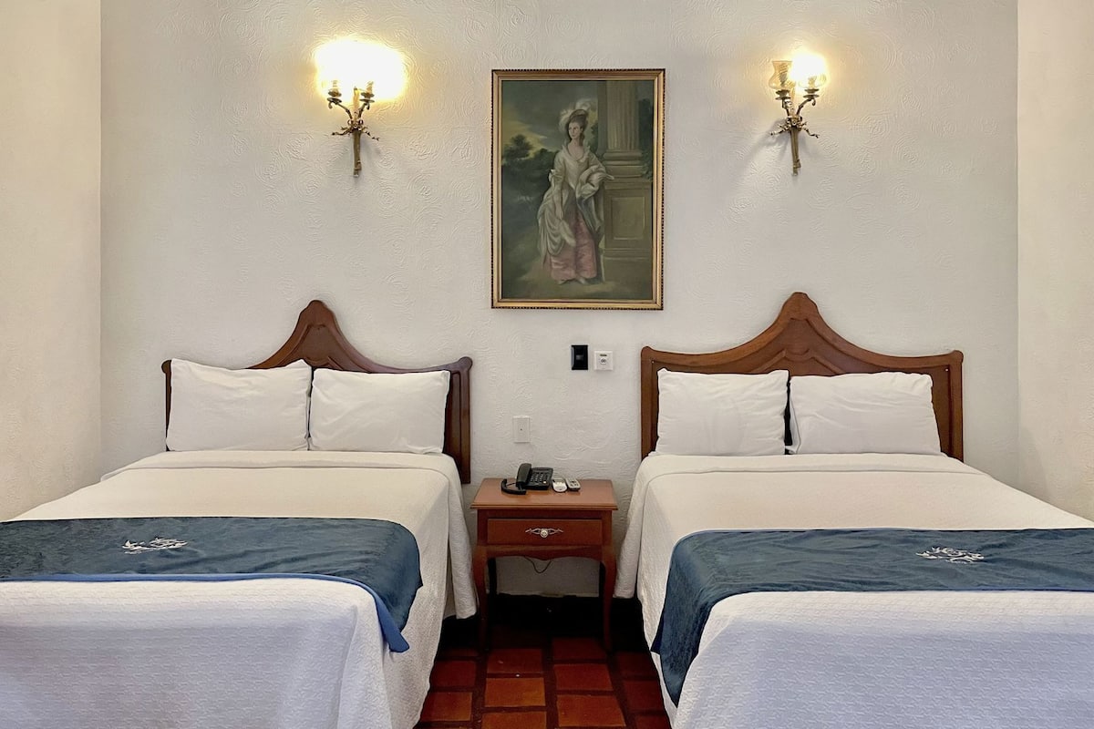 Una habitación de hotel con dos camas, una mesita de noche con teléfono e iluminación en la pared.