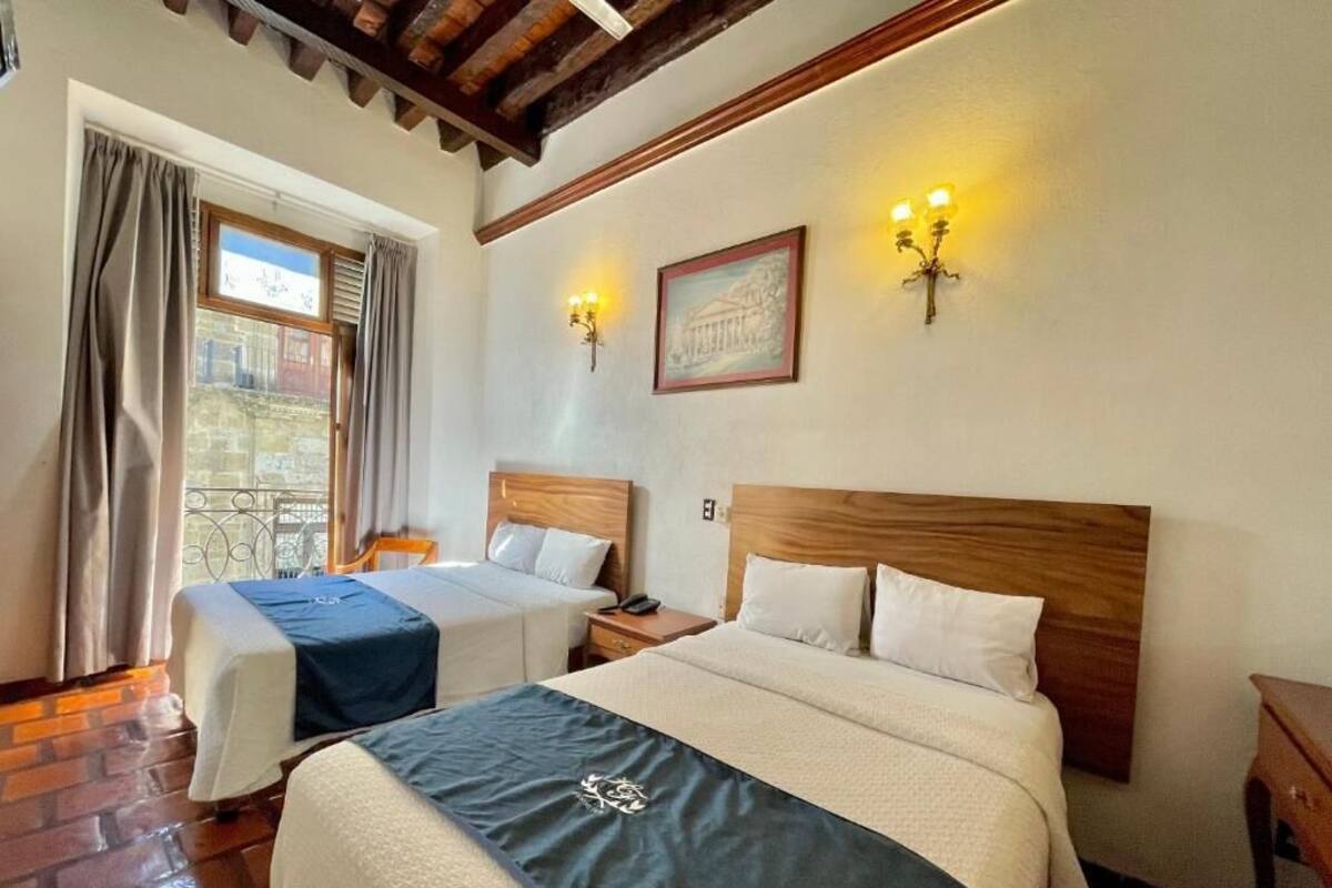 A hotel room with two beds, wooden headboards, a window with curtains, a framed picture on the wall, and wall-mounted lights.