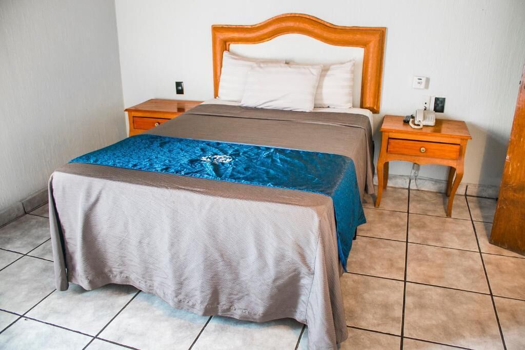 Un lit bien fait, avec une tête de lit en bois, une couvre-lit bleue et marron, et un plaid assorti au pied du lit. On y trouve deux tables de chevet en bois, dont l’une porte un téléphone.