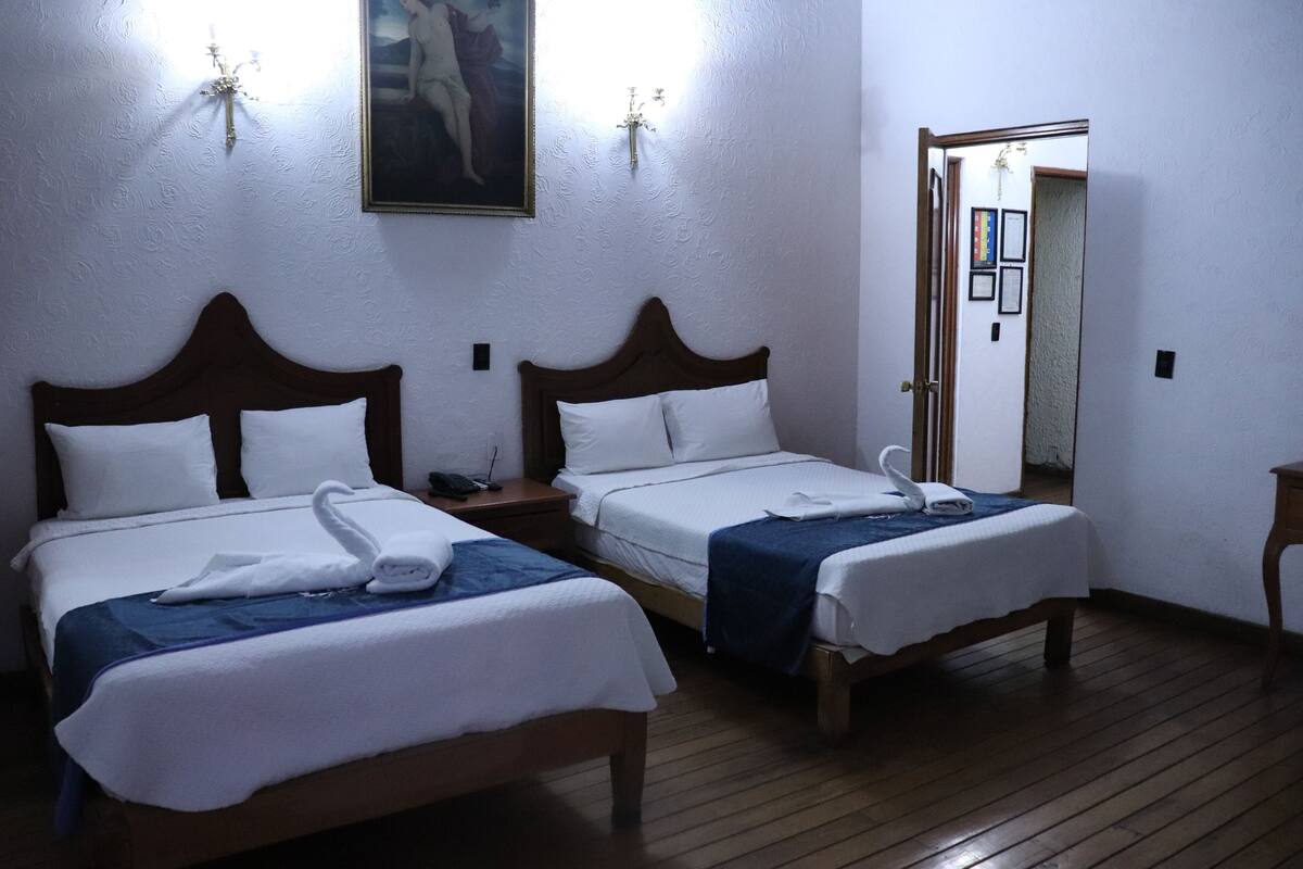 Two single beds with wooden frames and white linens, a painting on the wall, and a door leading to another room.