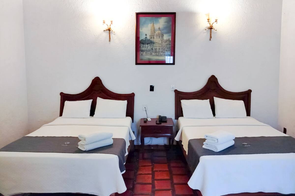 A hotel room with two beds, each with a wooden headboard, white bedding, and a folded towel on the bed. There is a nightstand with a phone and a lamp between the beds. A framed picture hangs on the wall above the beds.