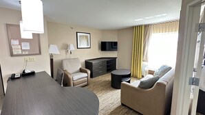 Ein Hotelzimmer mit Schreibtisch, zwei Sesseln, einer Couch, einem Flachbildfernseher, einer Lampe und einem fenster mit Vorhängen.