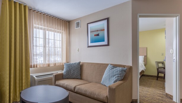 Ein Hotelzimmer mit beiger Couch, zwei Kissen, einem runden Tisch, einem Fenster mit Vorhängen, einem gerahmten Bild an der Wand und einer Tür zu einem anderen Raum.
