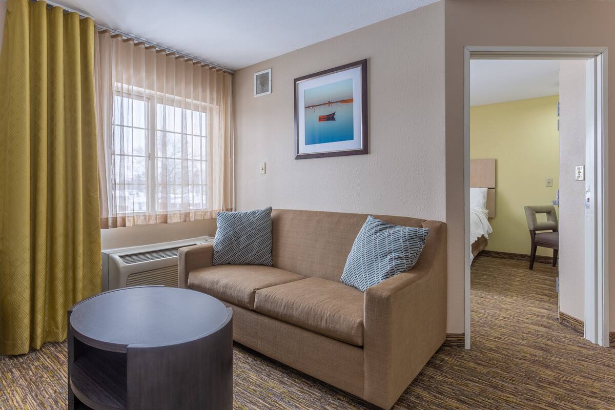 Une chambre d’hôtel avec un canapé beige, deux coussins, une table ronde, une fenêtre avec des rideaux, un tableau encadré au mur et une porte donnant sur une autre pièce.
