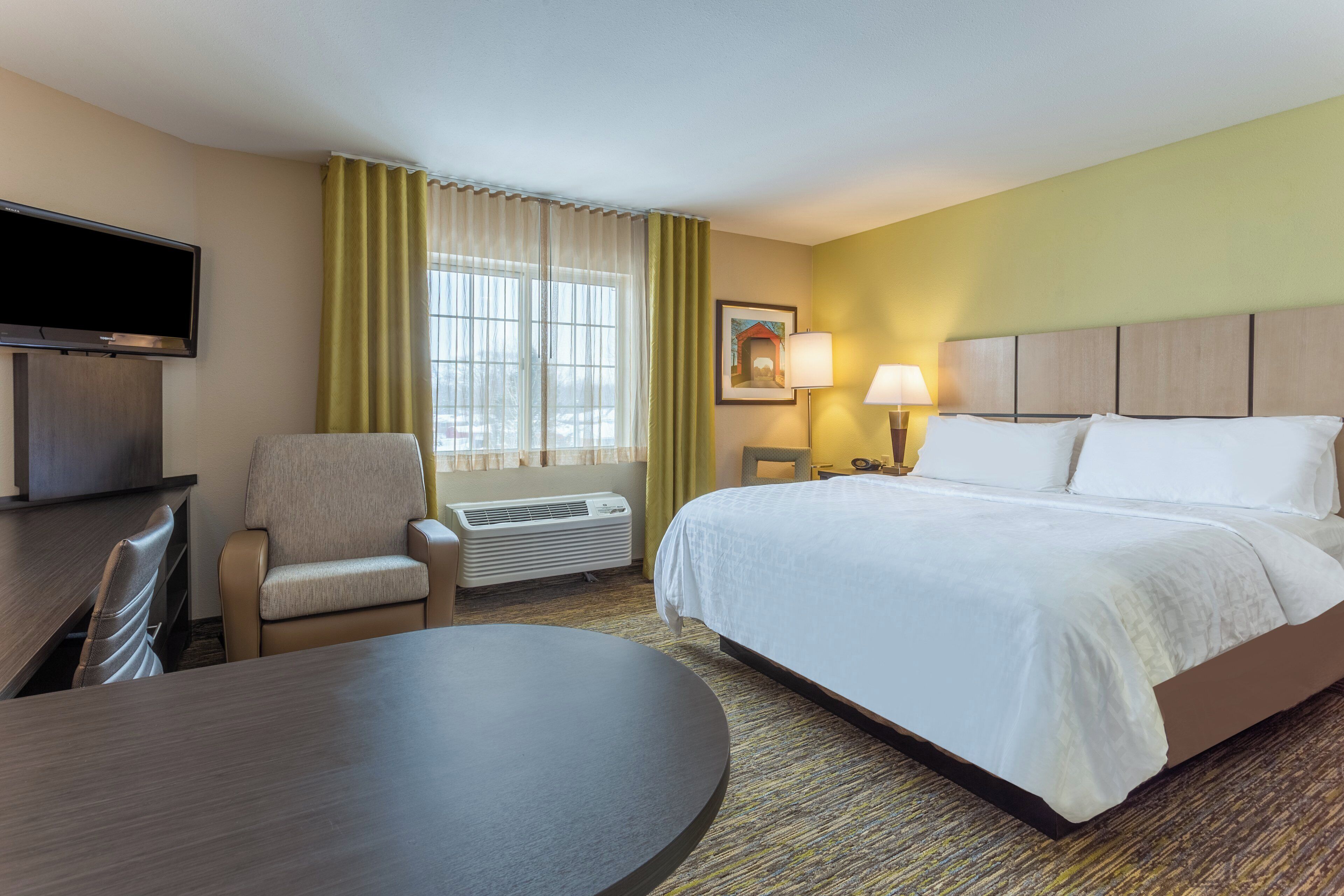 Quarto de hotel com cama, escrivaninha, cadeira, televisão, janela com cortinas, luminária e um quadro na parede.