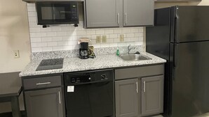 Eine Küche mit grauen Schränken, einem schwarzen Kühlschrank, einer Mikrowelle, einer Kaffeemaschine und einem Geschirrspüler.