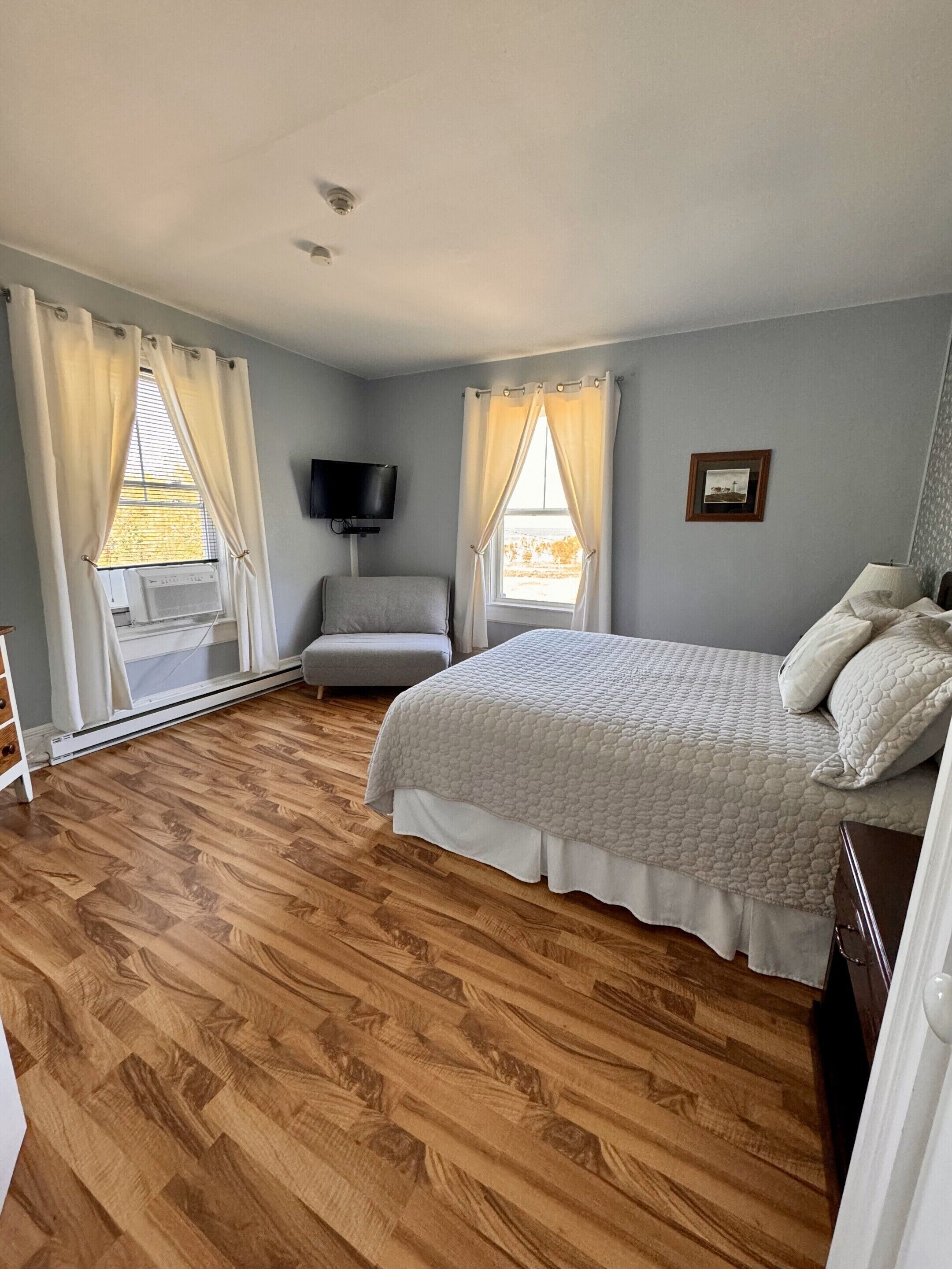 Un dormitorio con una cama, una silla, un televisor y una ventana con cortinas.