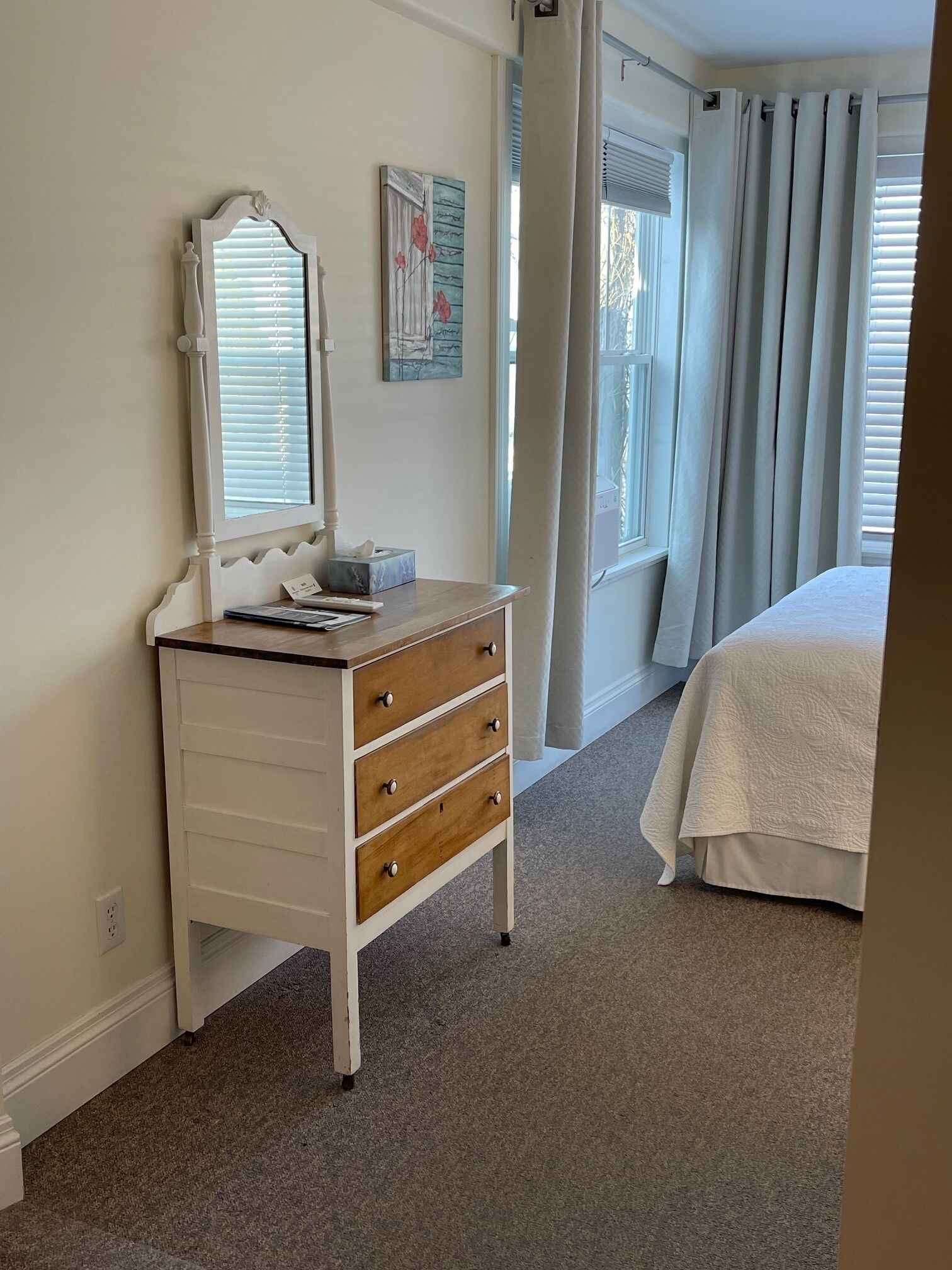 A bedroom with a dresser, a bed, a window with curtains, and a painting on the wall.