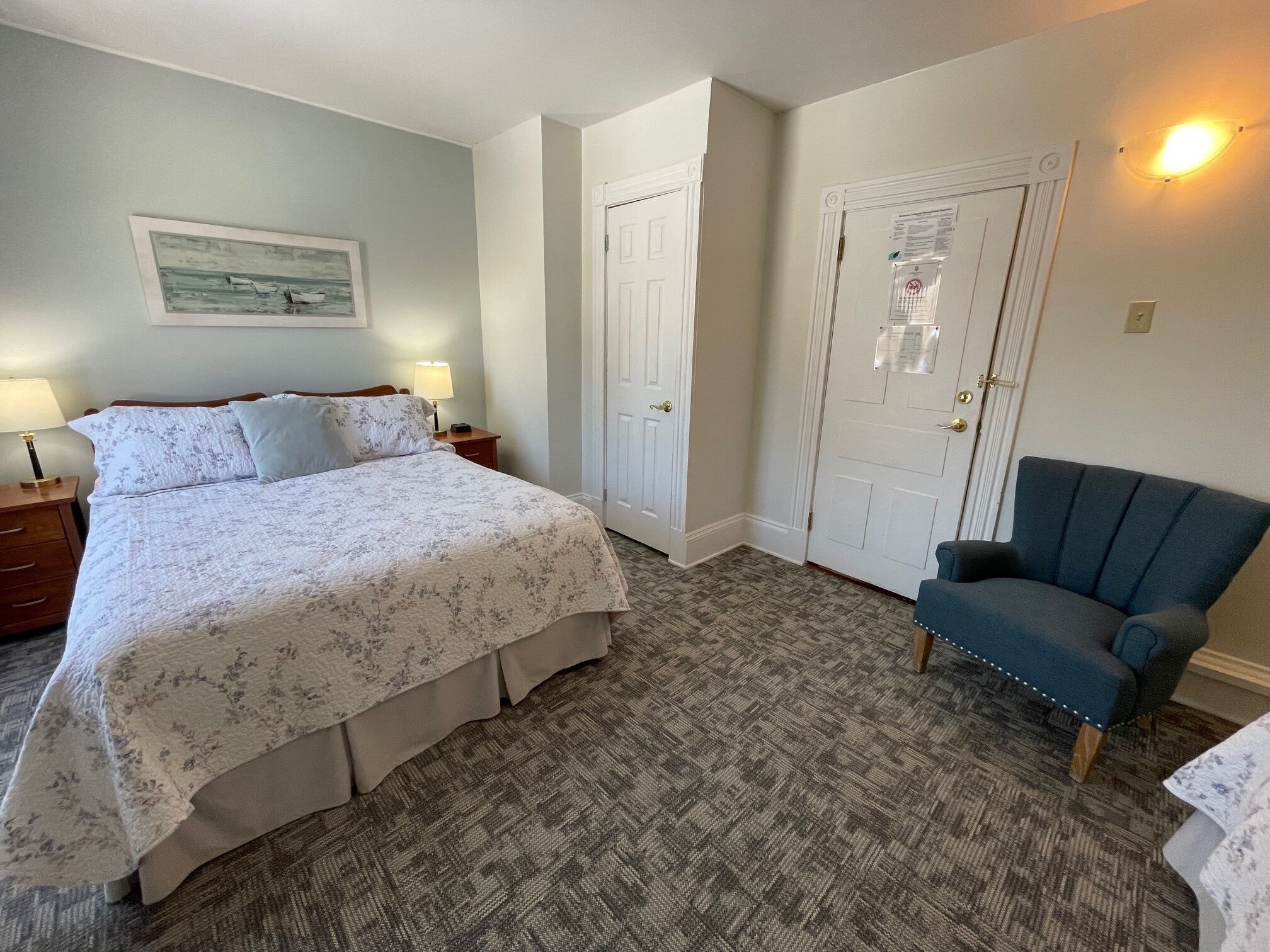 Une chambre à coucher avec un lit, deux tables de chevet avec des lampes, une chaise et une porte avec un interrupteur.