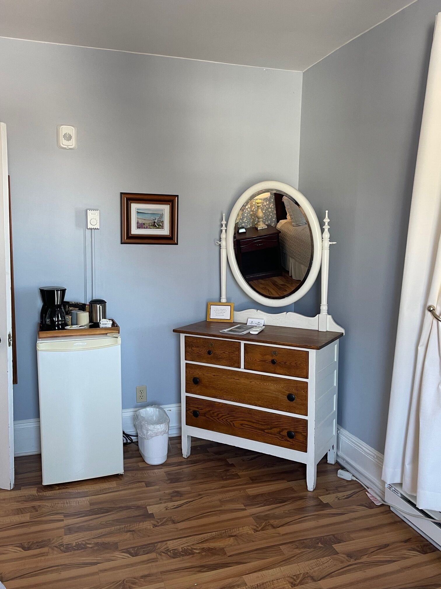 A bedroom with a dresser, a mirror, a framed picture, a coffee maker, and a trash bin.