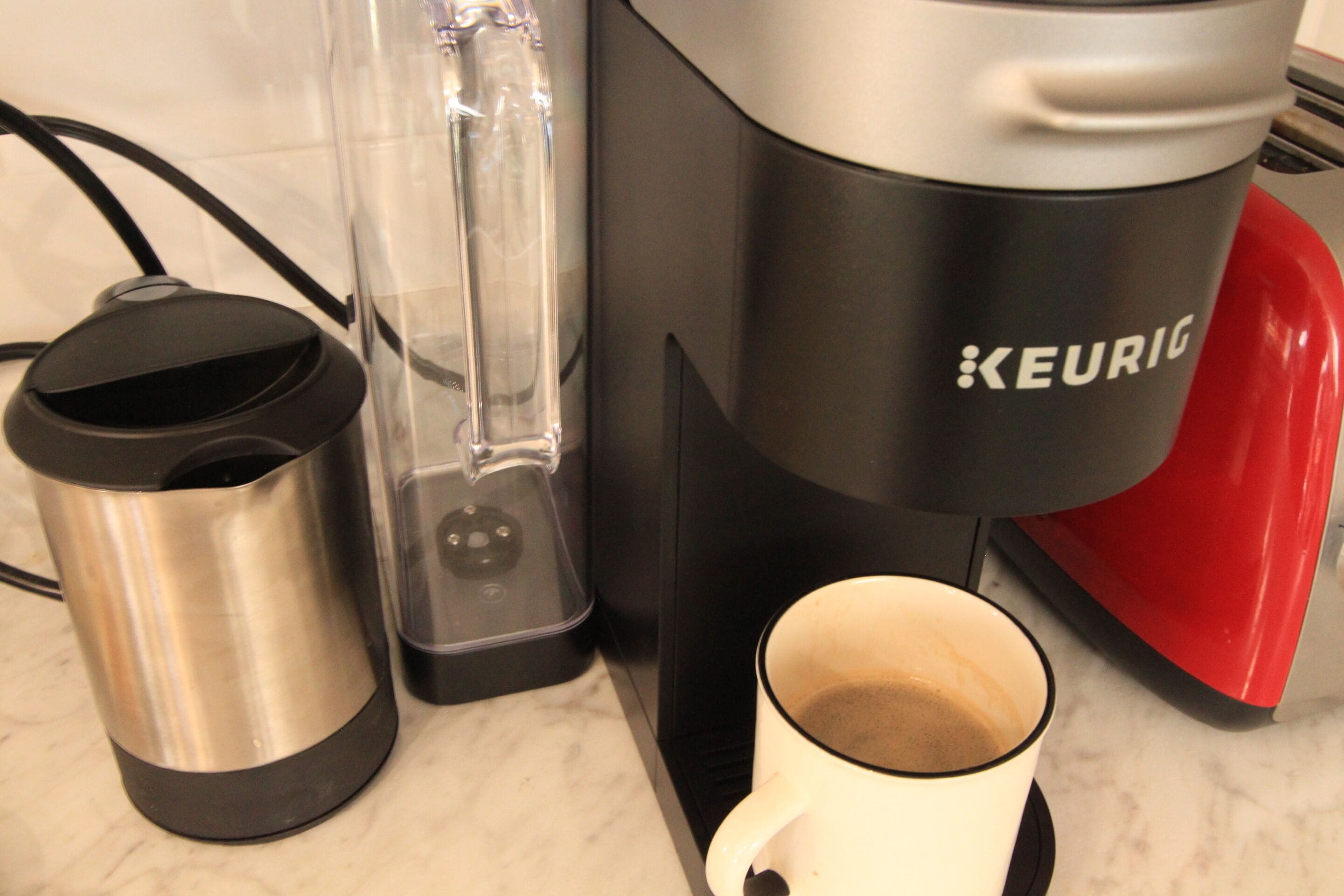A Keurig coffee maker, a stainless steel mug, and a white mug with coffee.