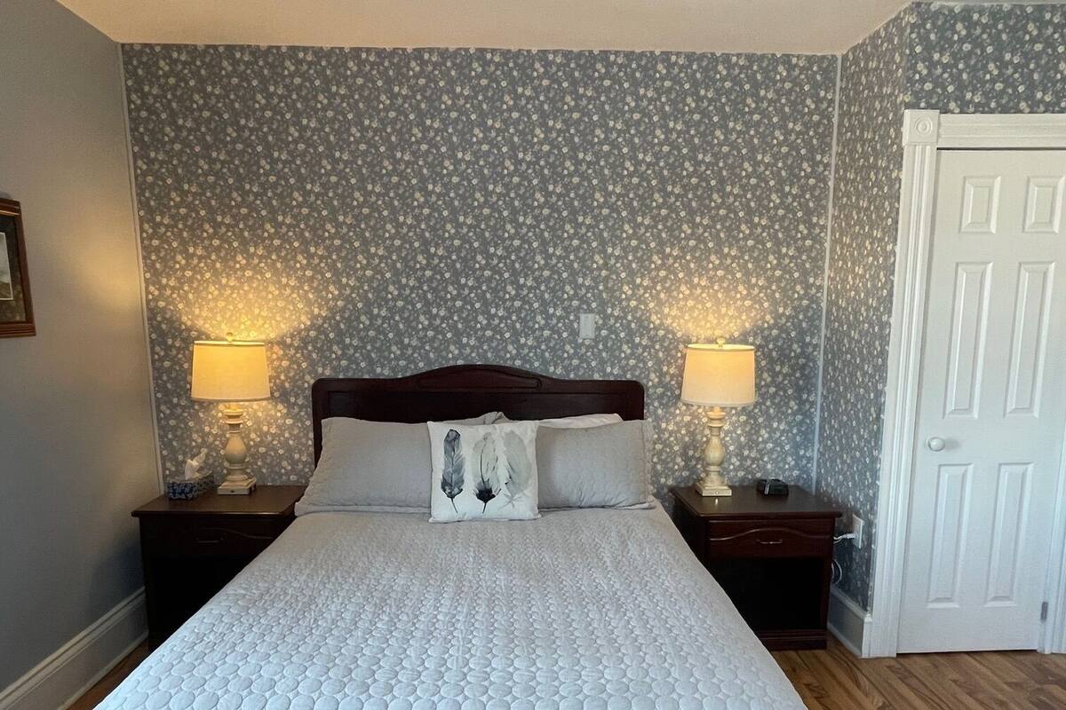 Una cama bien hecha con cubrecama acolchado, dos lámparas de noche, una mesita de madera y una puerta a la derecha.