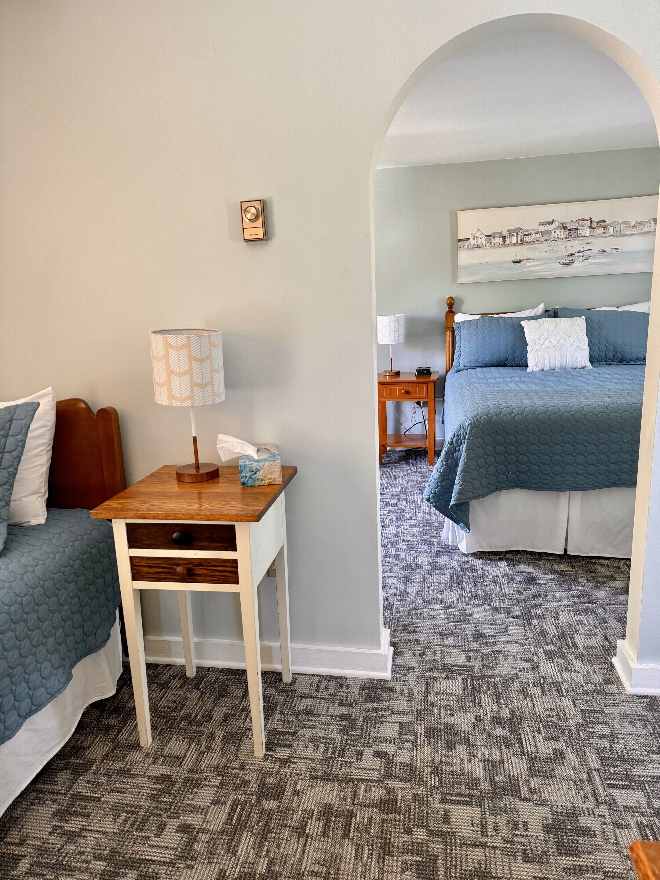 A hotel room with a bed, a desk, a lamp, a clock, and a painting on the wall.