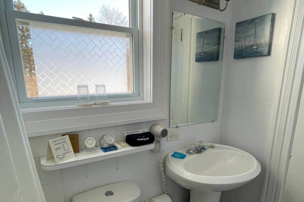 Un baño con inodoro, bidé, lavamanos y ventana con vista a árboles y una valla.