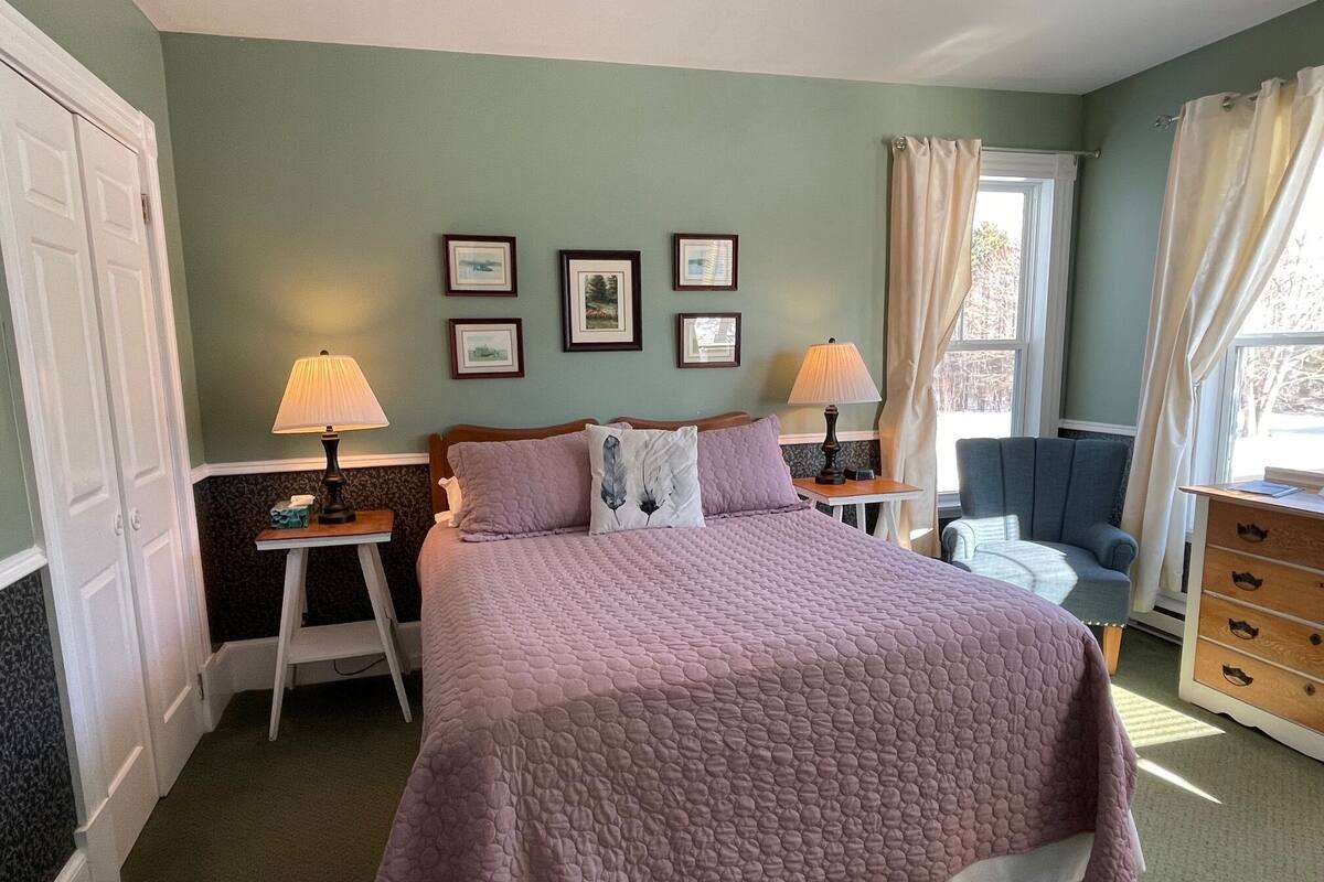 Un dormitorio con cama, dos mesitas de noche con lámparas, cómoda, silla y ventana con cortinas.