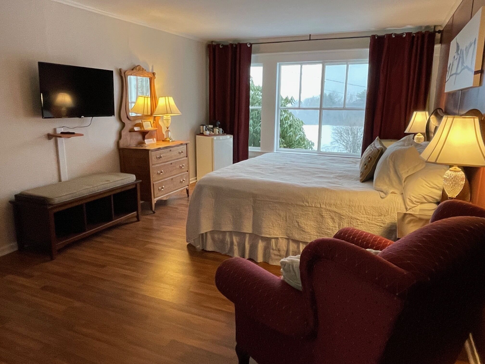 Un dormitorio con cama, lámparas de noche, televisor, cómoda, banca y ventana con cortinas.