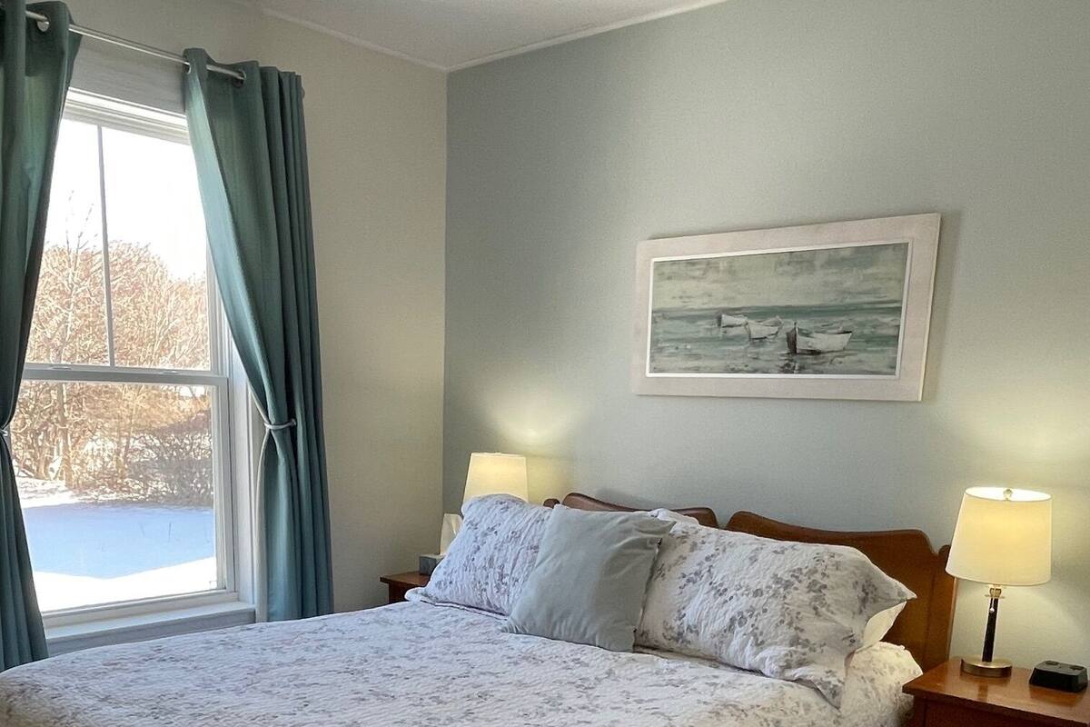 Un dormitorio con una cama grande, una mesita de noche con lámpara, un cuadro en la pared y una ventana con cortinas.