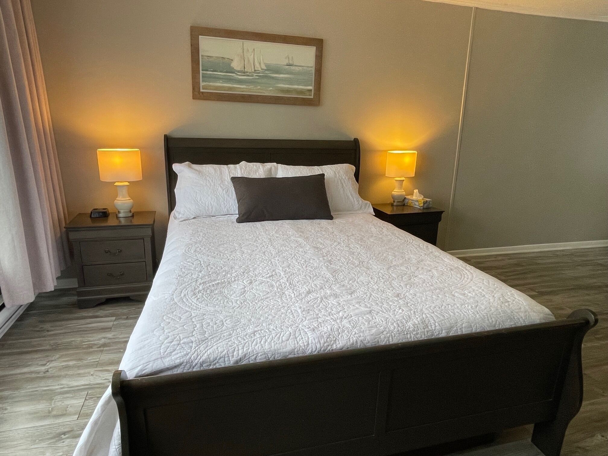 A neatly made bed with white bedding, two bedside lamps, a nightstand with a clock, a framed picture on the wall, and a wooden floor.