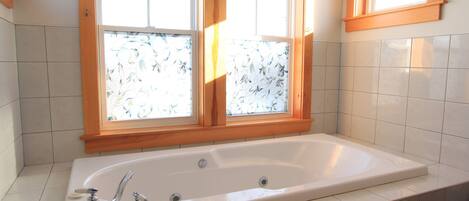 Un baño con una bañera grande, dos ventanas con vidrio esmerilado y paredes de azulejos blancos.
