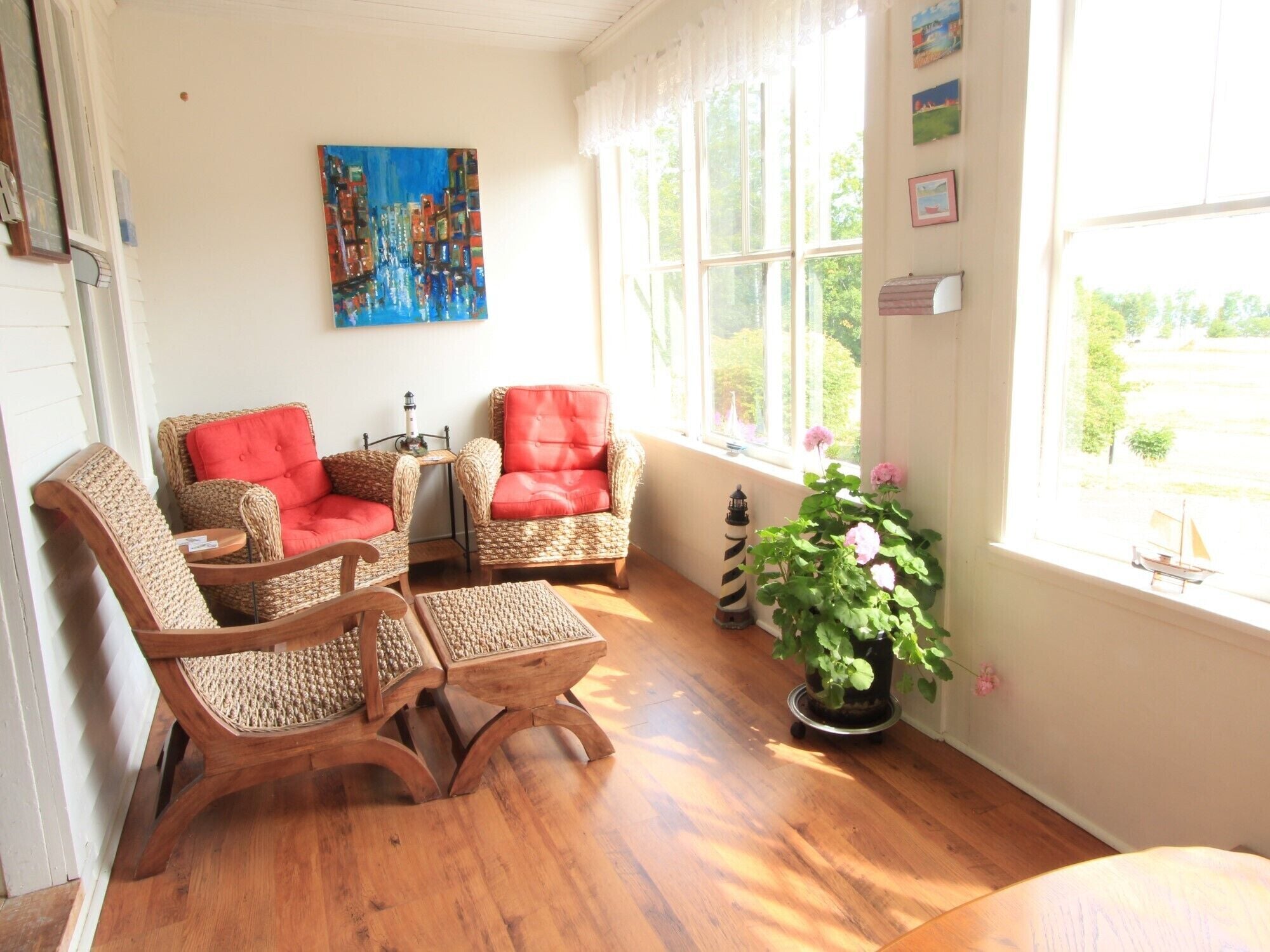 A room with wicker chairs, a small table, a painting, a potted plant, and a view through the window.