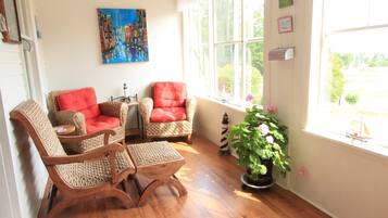 A room with wicker chairs, a small table, a painting, a potted plant, and a view through the window.