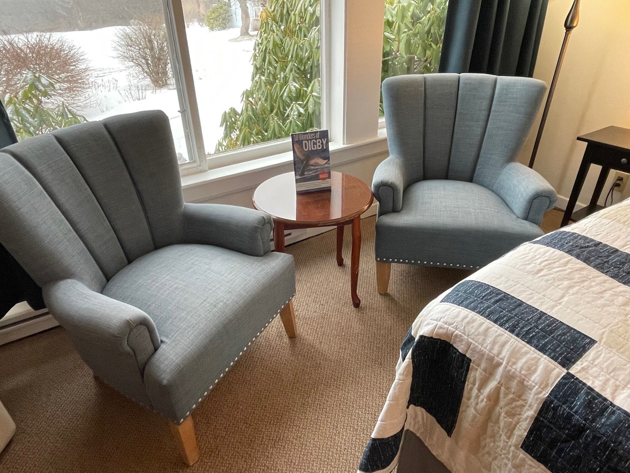 A hotel room with two grey armchairs, a round wooden table, and a bed with a quilt.