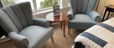 A hotel room with two grey armchairs, a round wooden table, and a bed with a quilt.