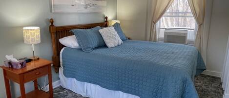 Un dormitorio con cama, mesita de noche, lámpara, un cuadro y ventana con cortinas.