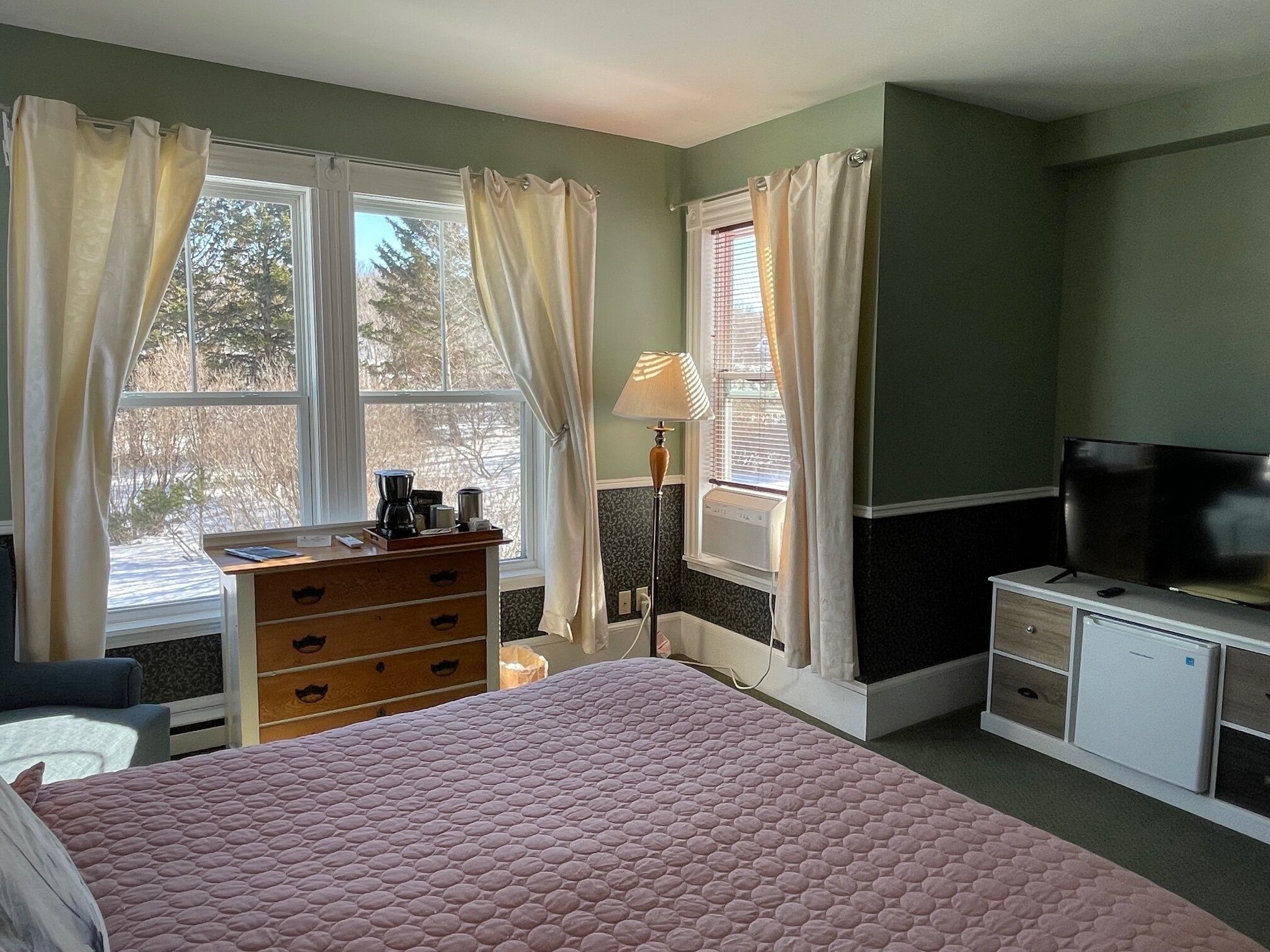 A bedroom with a bed, dresser, television, and windows with curtains.