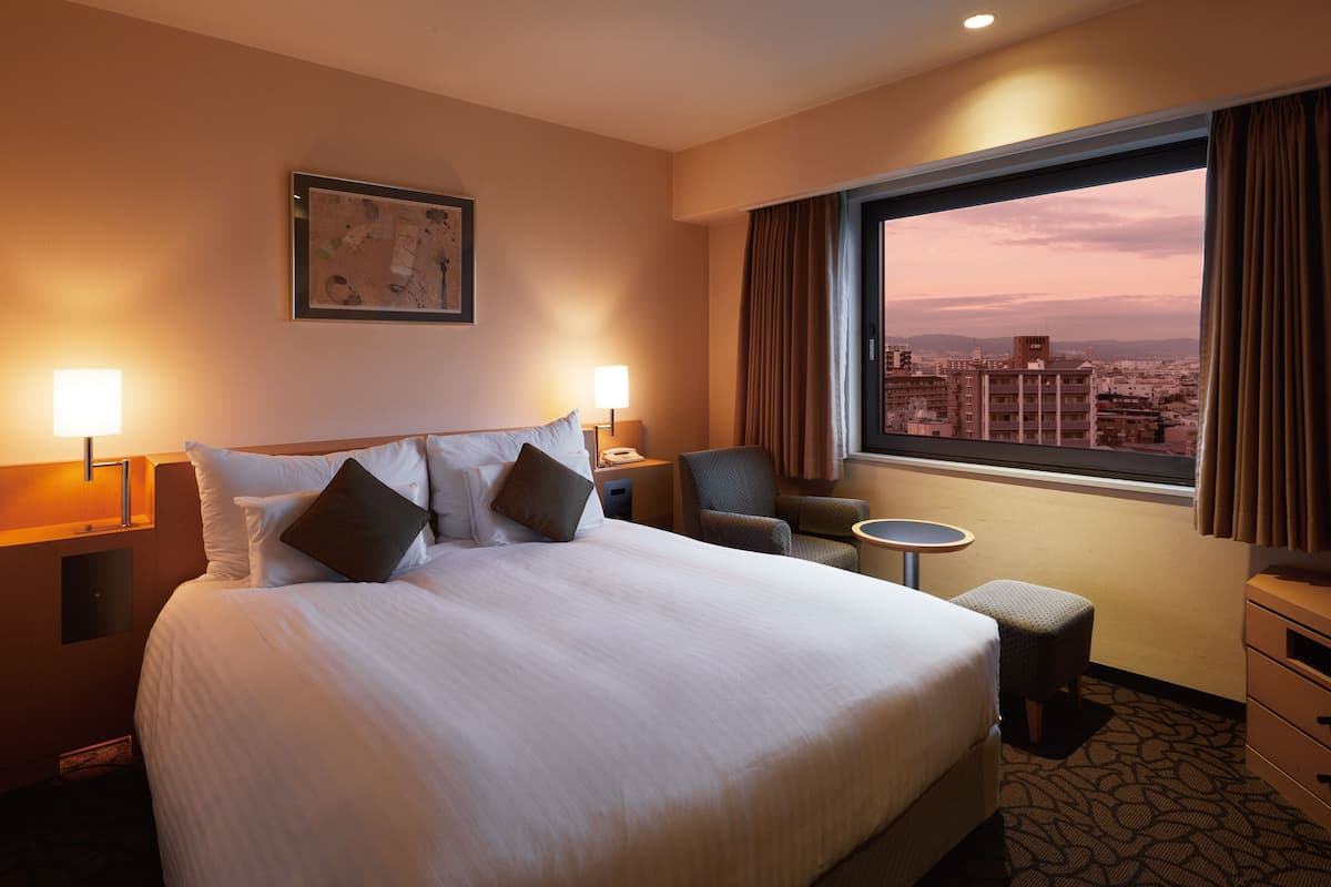 Habitación de hotel con una cama grande, dos lámparas de noche, una silla, una mesita y una ventana con vistas a la ciudad.