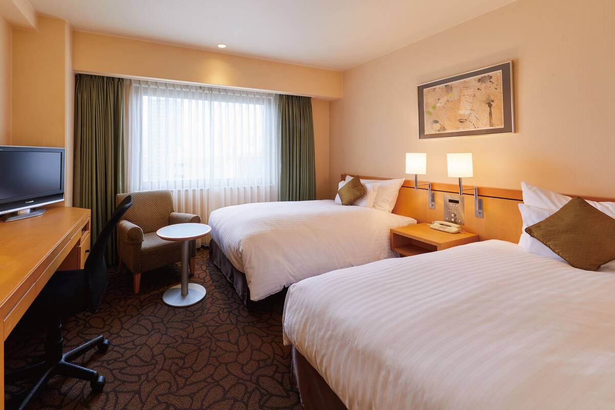 Una habitación de hotel con dos camas, un escritorio con televisor, una silla, dos mesitas de noche con lámparas y un ventanal con cortinas.