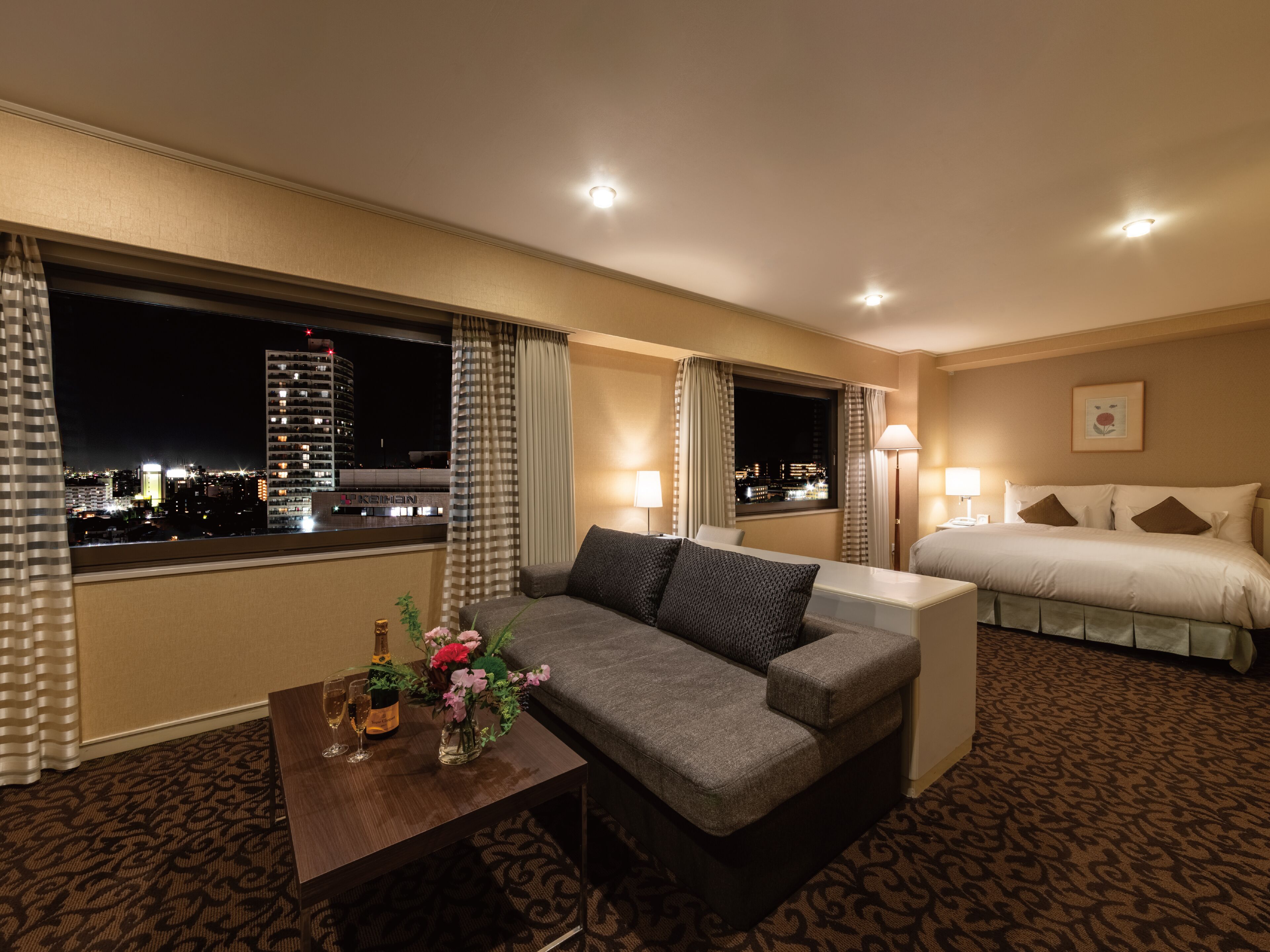 A hotel room with a sofa, a bed, a nightstand, a lamp, and a view of the cityscape.