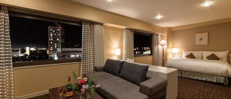 A hotel room with a sofa, a bed, a nightstand, a lamp, and a view of the cityscape.