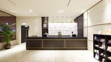 A reception area with a dark counter, two receptionists, a potted plant, and a bookshelf with brochures.