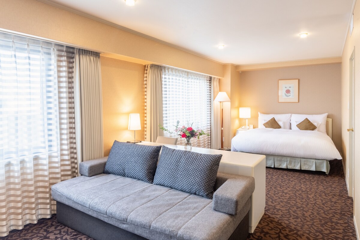 Une chambre d’hôtel avec un canapé, un lit, une table de chevet avec des lampes et une fenêtre avec des rideaux.