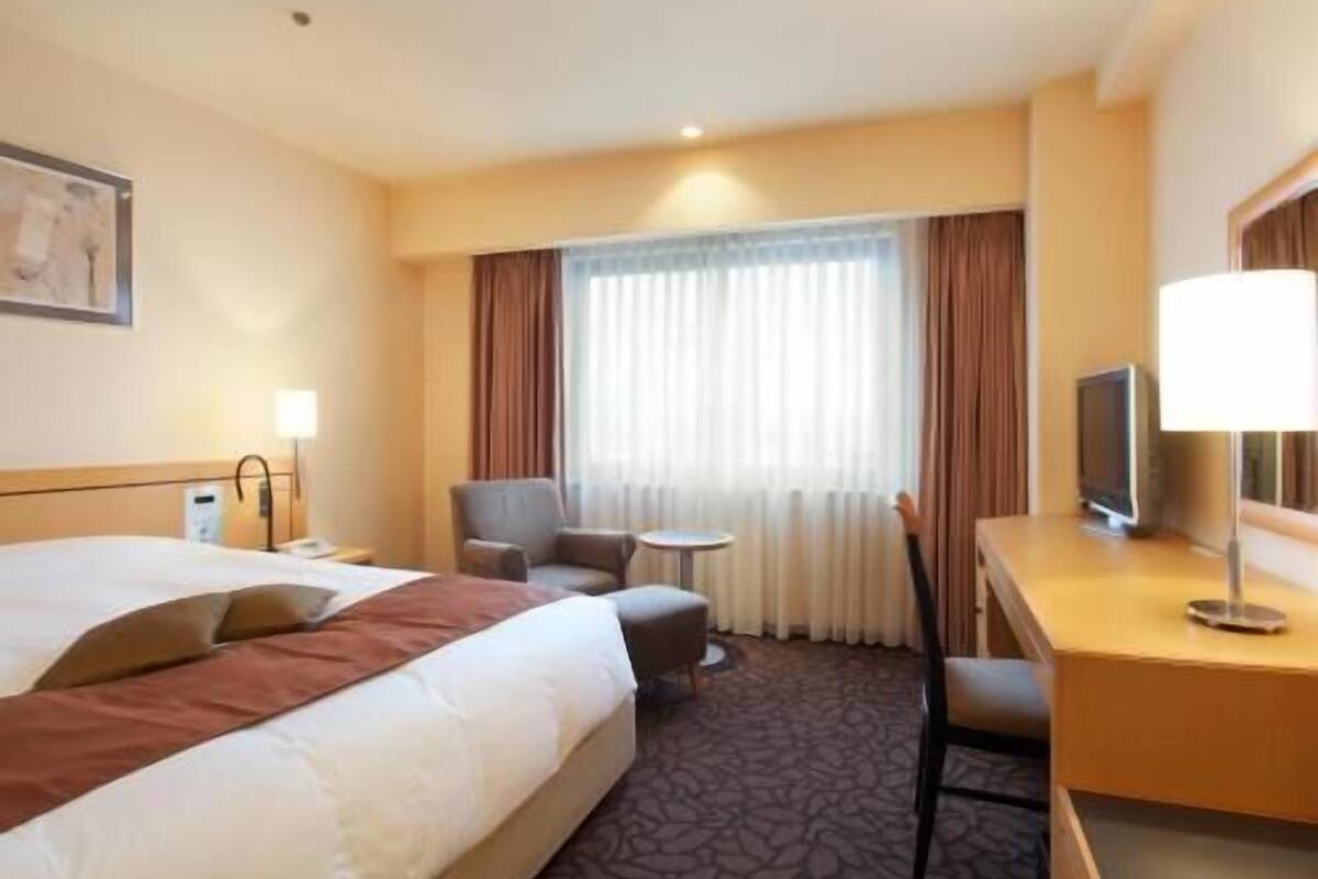 Habitación de hotel con cama, escritorio, silla, televisor y ventana con cortinas.