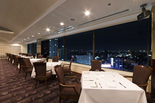 Comedor de hotel con mesas dispuestas para el servicio, amplias ventanas con vista a la ciudad de noche.