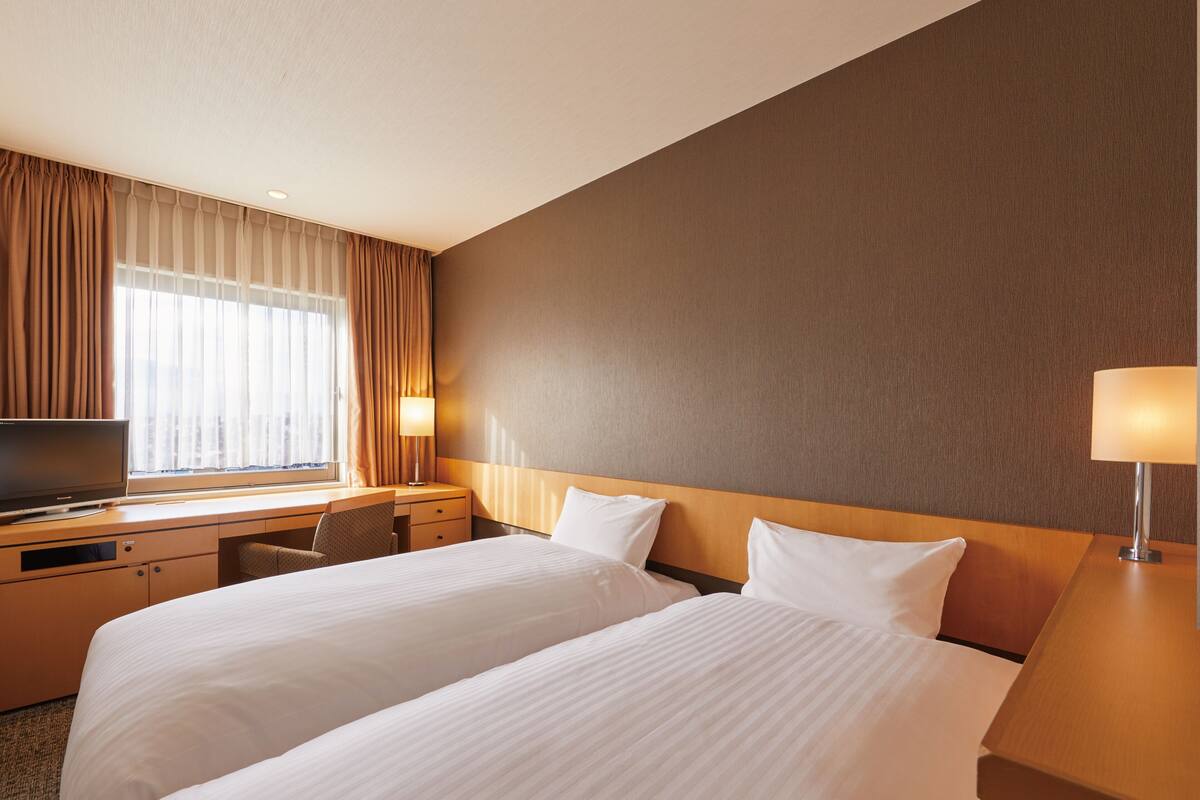 Habitación de hotel con dos camas, un escritorio con televisor y un ventanal con cortinas.