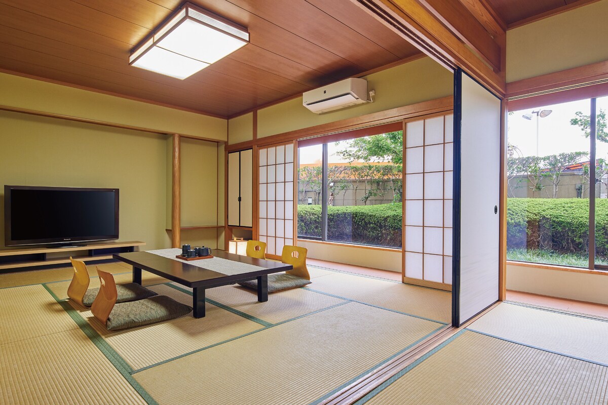 Une pièce de style japonais traditionnel, avec un sol recouvert de tatamis, un téléviseur et une porte coulissante donnant sur un paysage verdoyant.