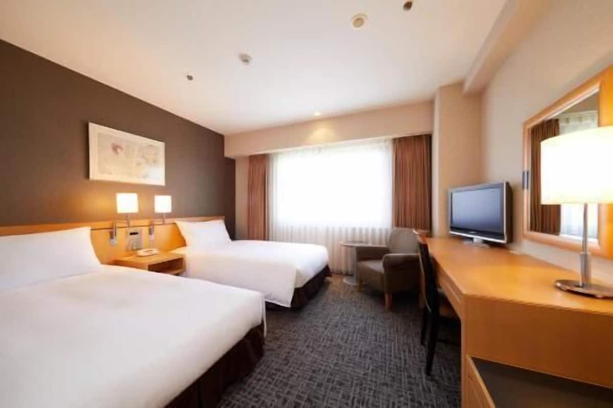 Habitación de hotel con dos camas, un escritorio, televisor, una silla y ventana con cortinas.
