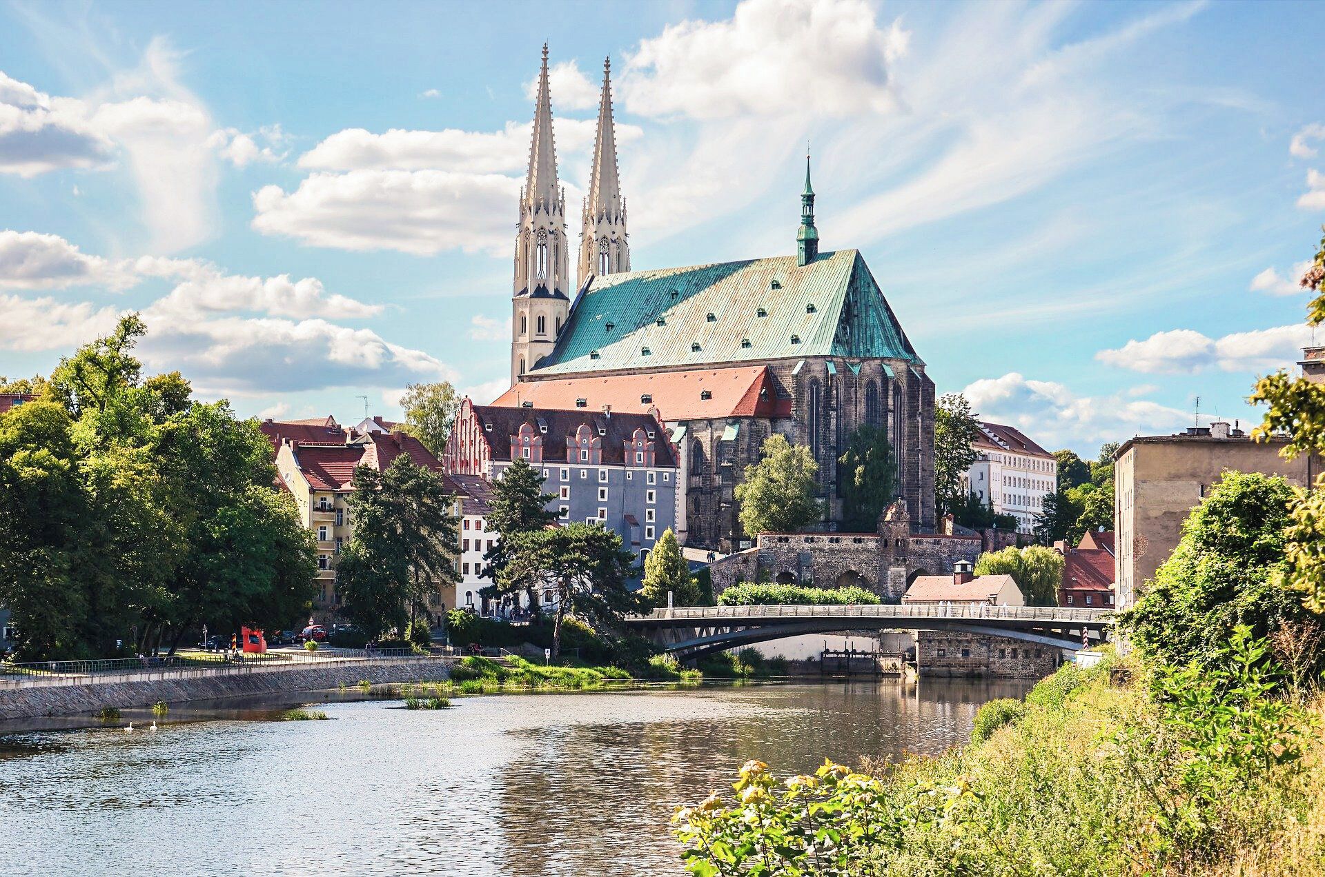 A historic church with twin spires and a green roof, surrounded by trees and a river.