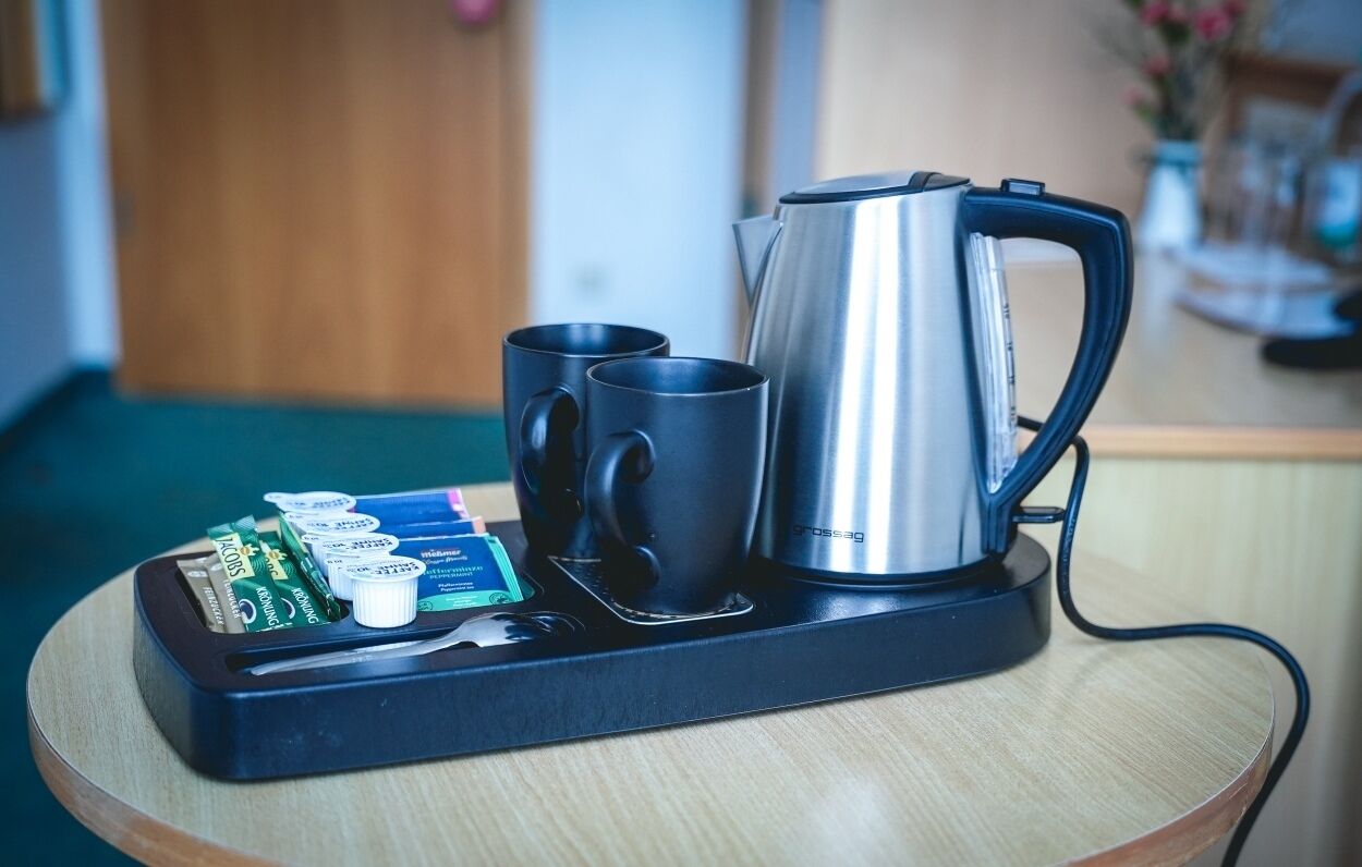 A coffee maker, two black mugs, and a tray with coffee packets on a wooden table.