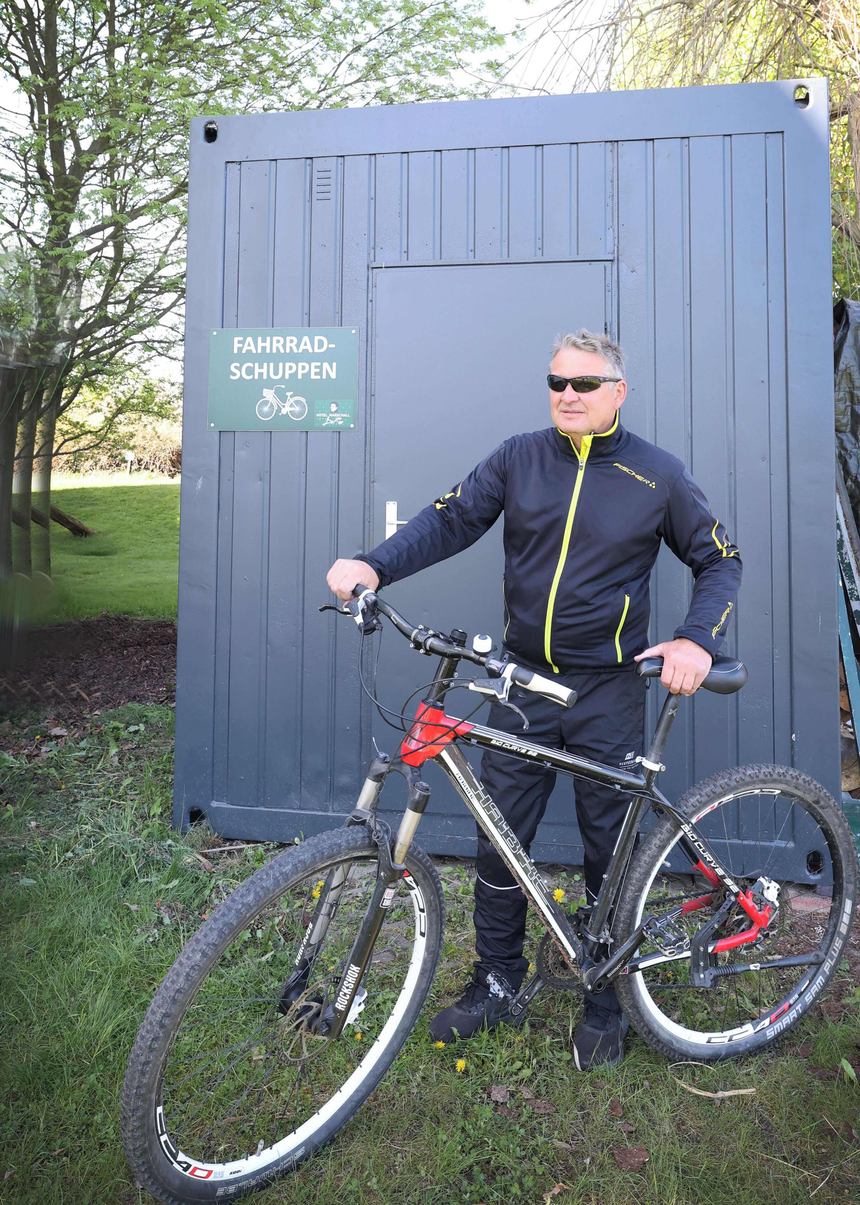 A man standing next to a bicycle in front of a grey structure with a sign that reads "Fahrrad-Schuppen".