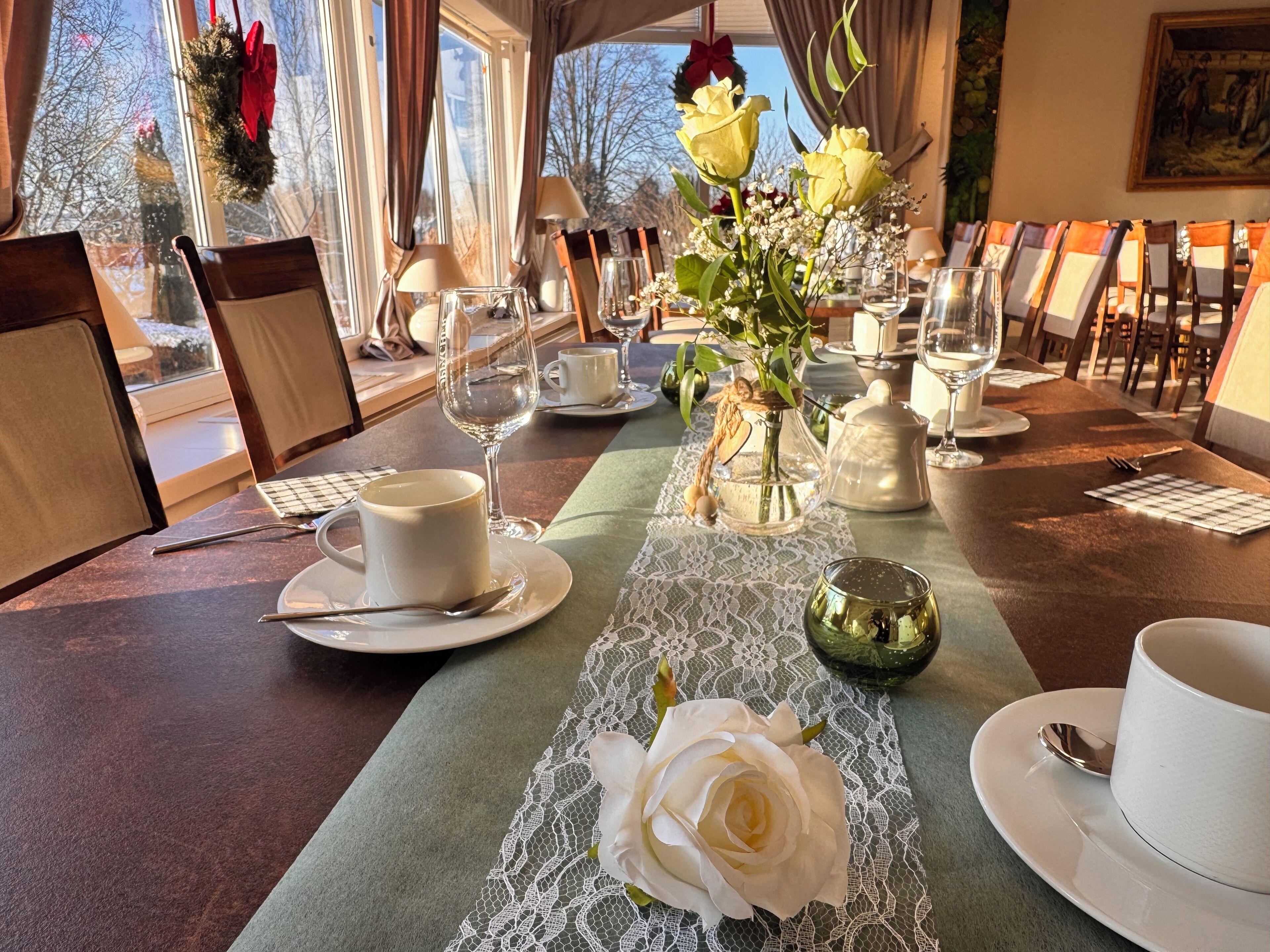 A dining table set with white cups, saucers, and a green glass bowl, with a lace table runner and a white rose centerpiece.