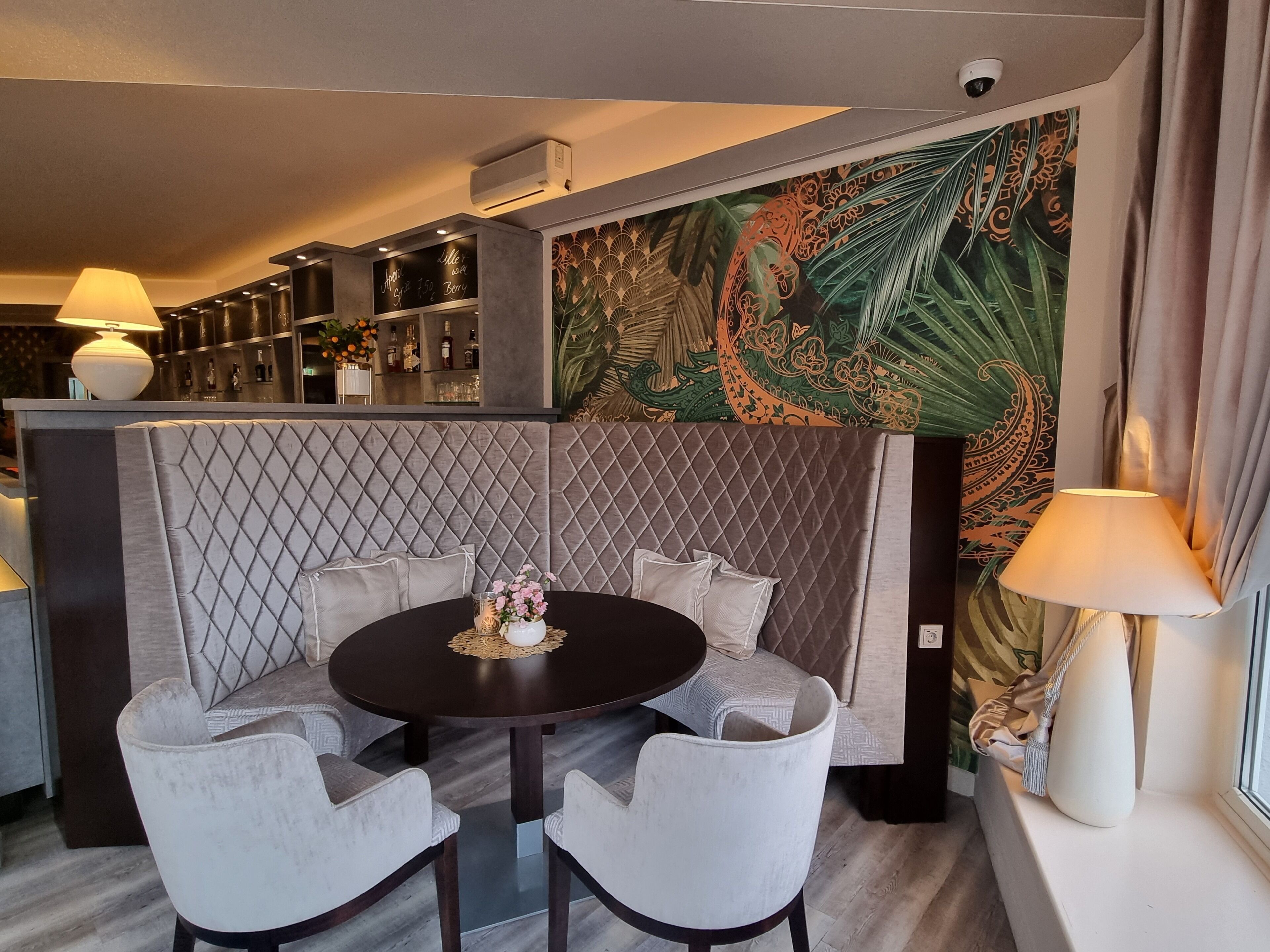 Un comedor moderno con una mesa redonda, sillas acolchadas y un centro de mesa decorativo. Se ve un área de bar con estantes llenos de botellas y un menú en una pizarra al fondo.