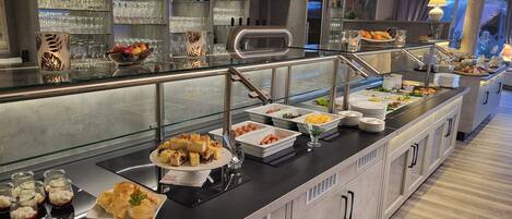 Un área de bufé con diversas opciones de comida, un bar con botellas de licores y vistas al exterior a través de amplias ventanas.
