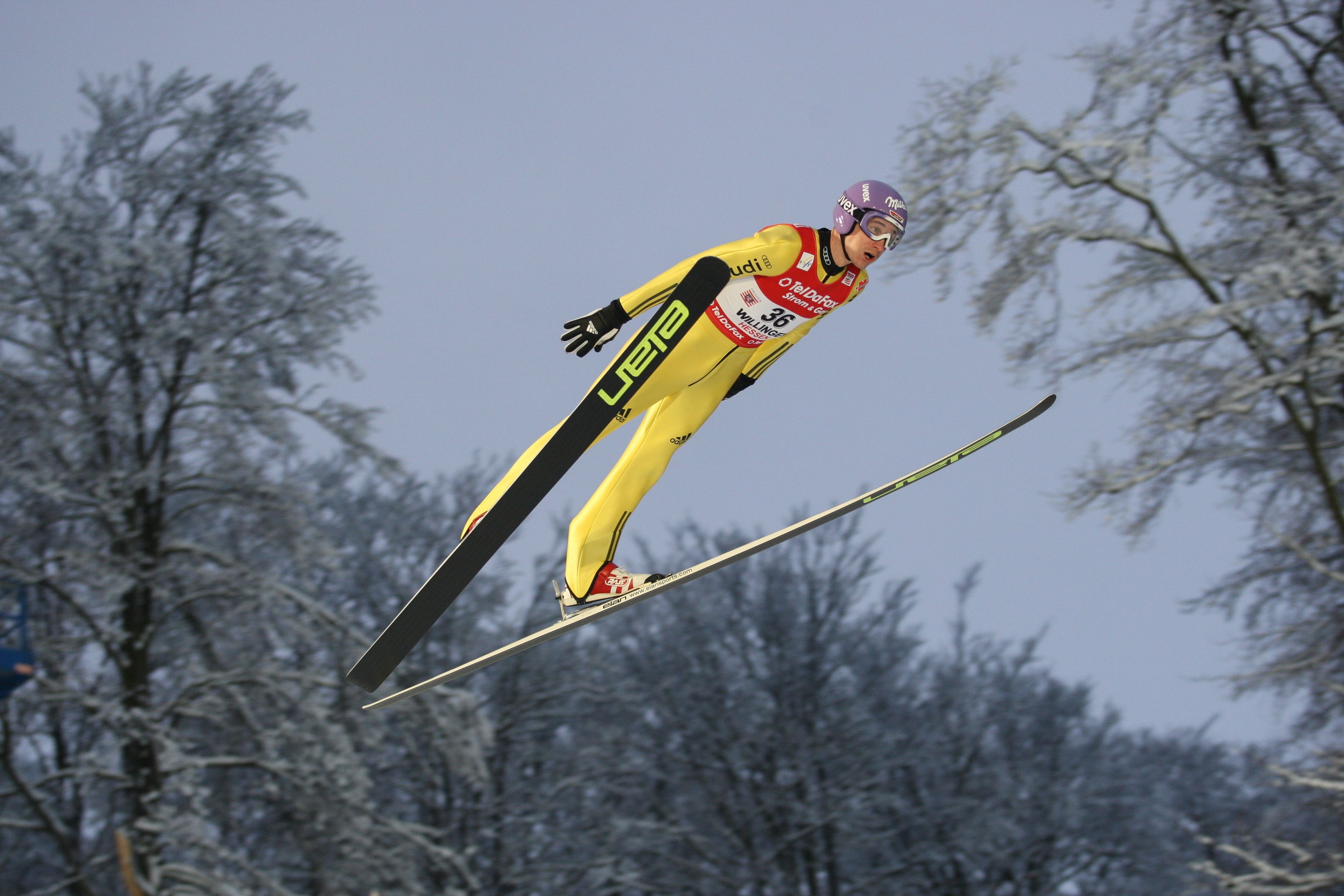 A skier in a yellow suit mid-air, with trees in the background.