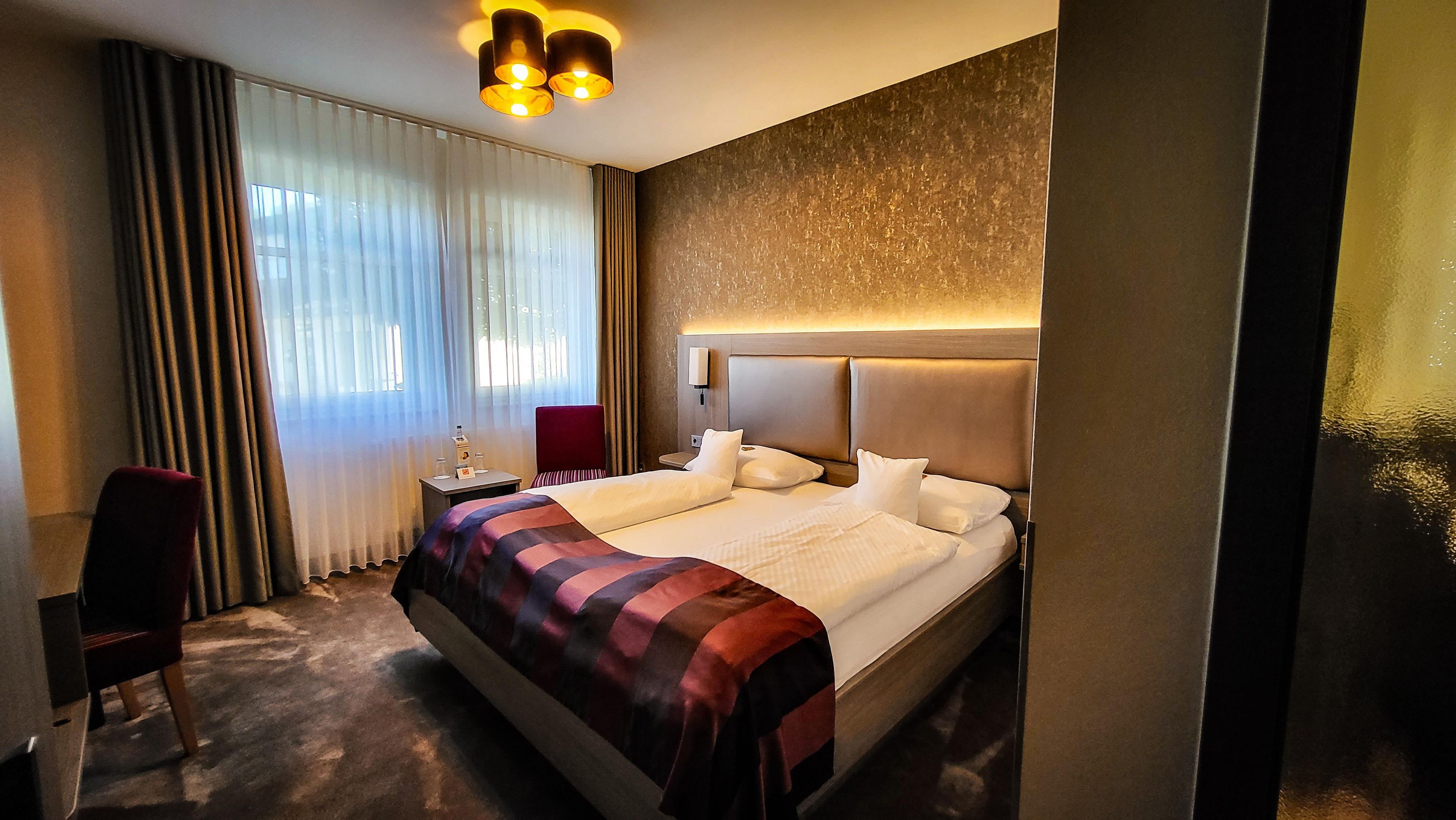 Ett hotellrum med en stor säng, ett skrivbord, en stol, ett nattduksbord och ett fönster med gardiner.
