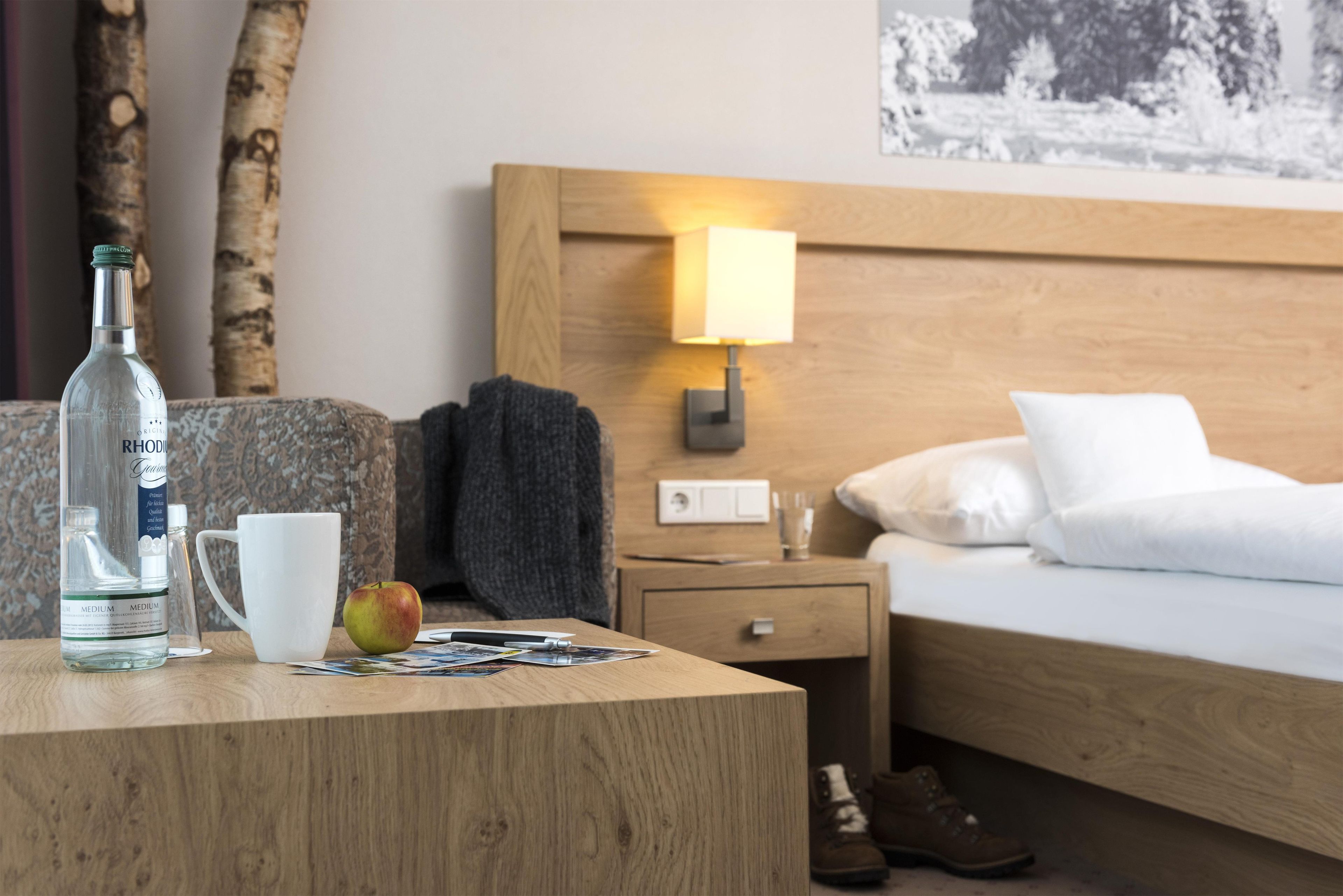 A hotel room with a wooden headboard, a bedside lamp, a bed with white bedding, a nightstand with a lamp, a wooden table with a bottle, a mug, and an apple.